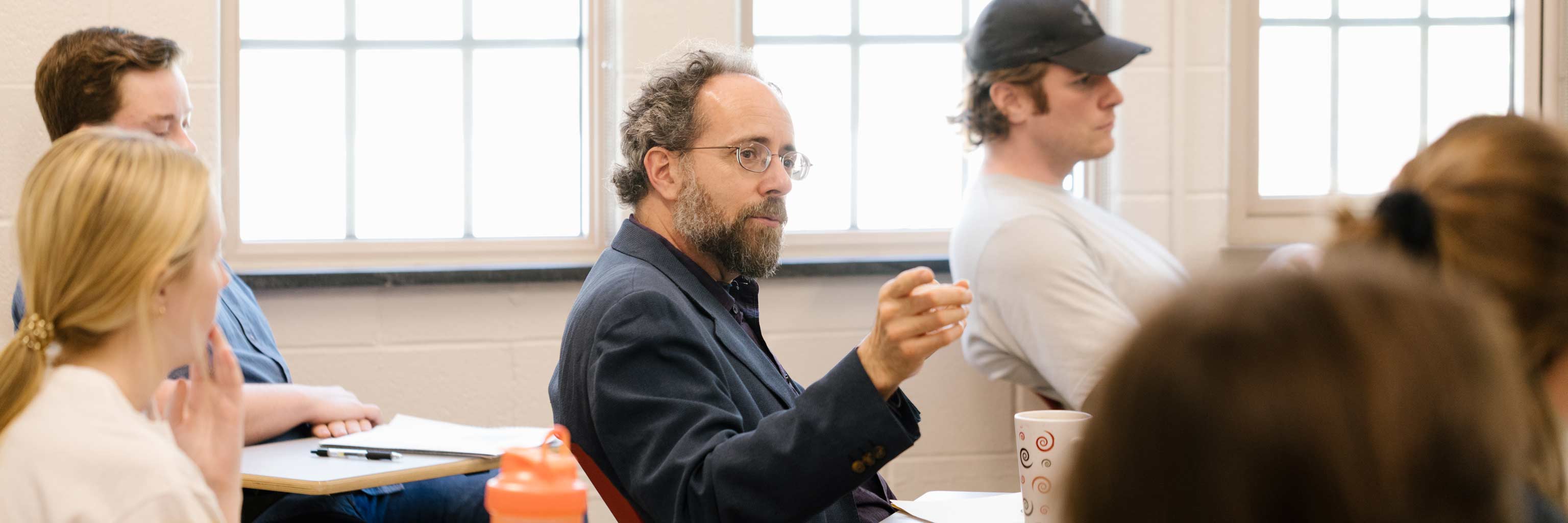 A professor speaks while sitting at a desk among a class of students.