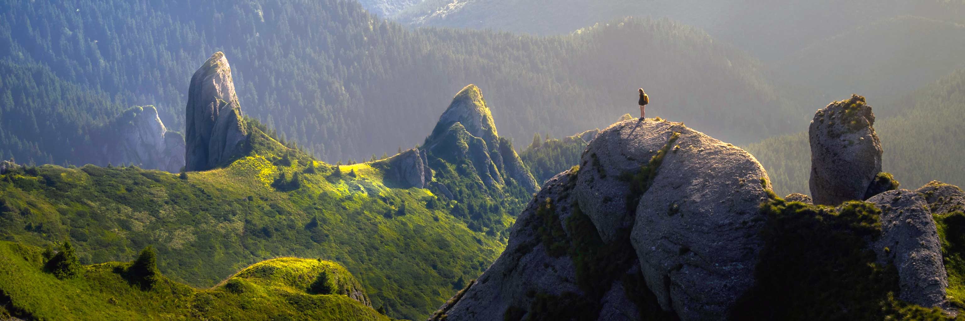A distant figure standing on top of a mountain peak amongst a beautiful green elevated landscape.