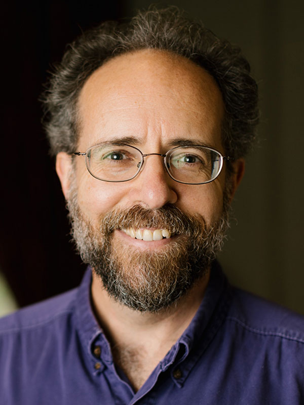 A headshot of Adam Leite, who wears a dark blue polo shirt and poses against a black background.