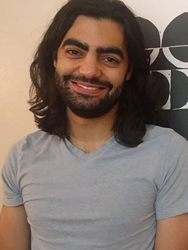 A headshot of Ricky Mouser, who wears a light blue v-neck shirt and poses against a wall.