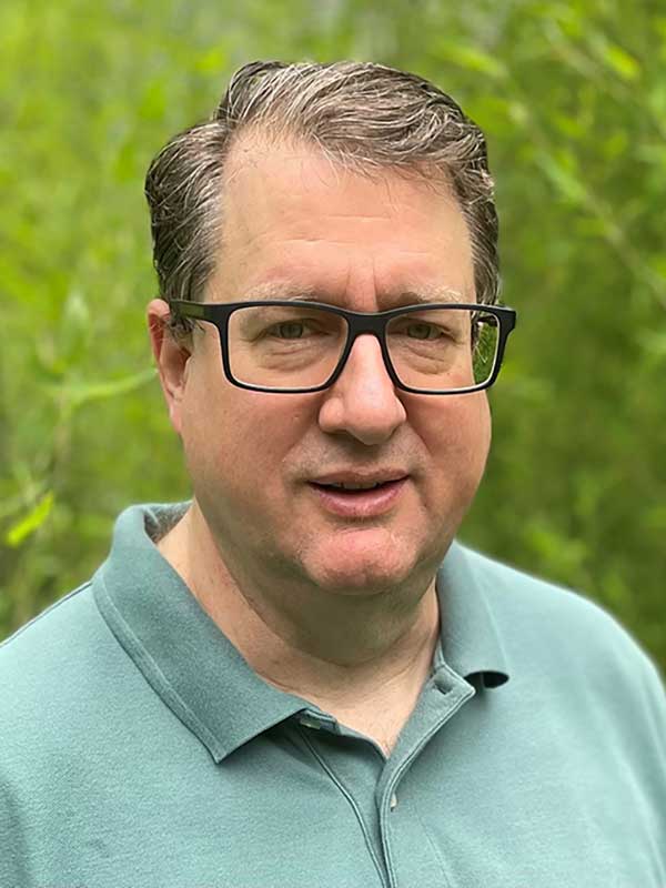 A headshot of David Sussman, who wears a green polo shirt and poses outside.